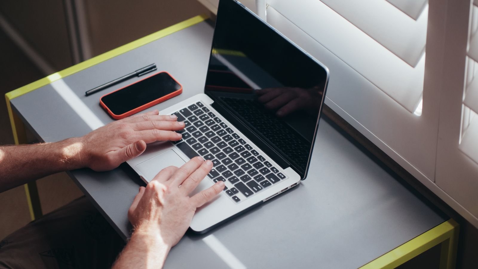 Hands typing on a laptop beside a window with a notebook and pen on the desk