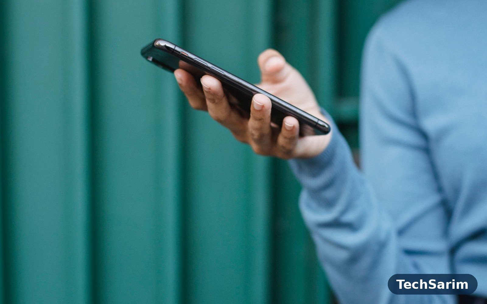 A person in a blue sweater holding a modern smartphone against a teal background