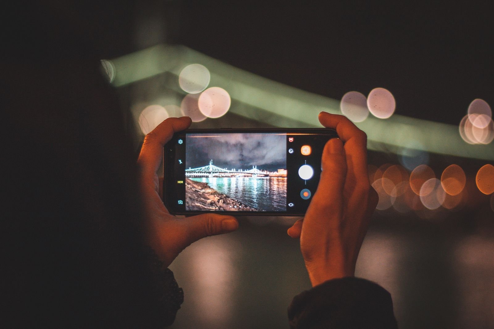 Hands holding a smartphone horizontally to photograph a lit bridge at night