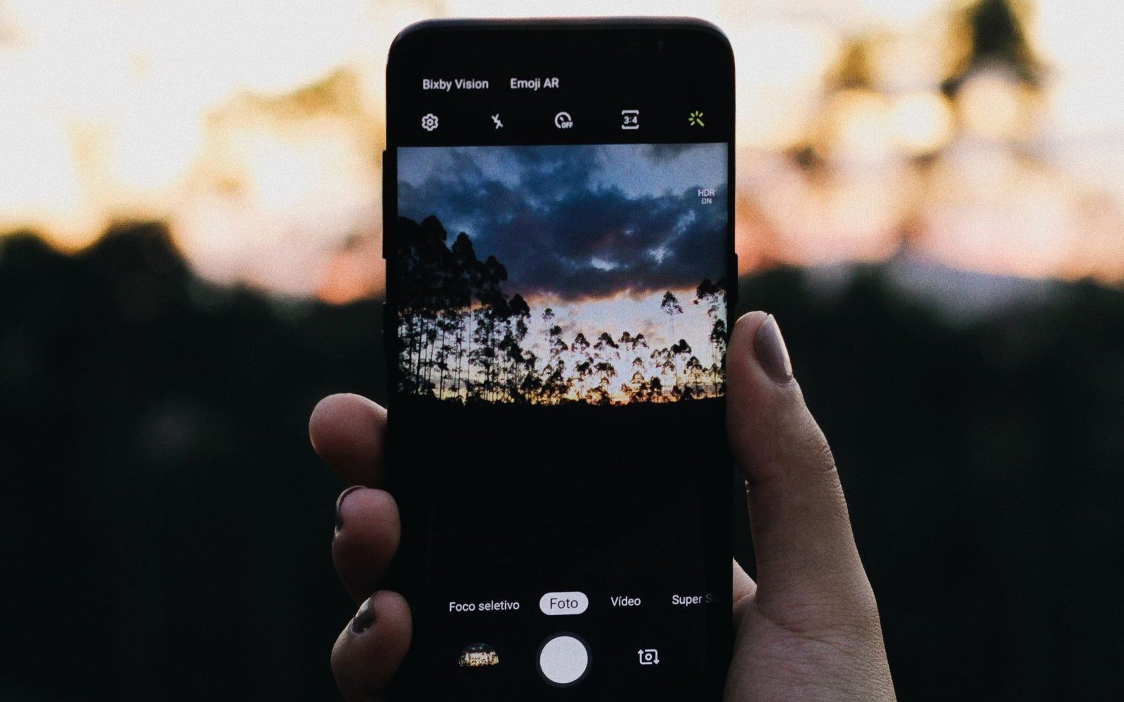 A hand holding a smartphone framing a forest sunset in the camera app
