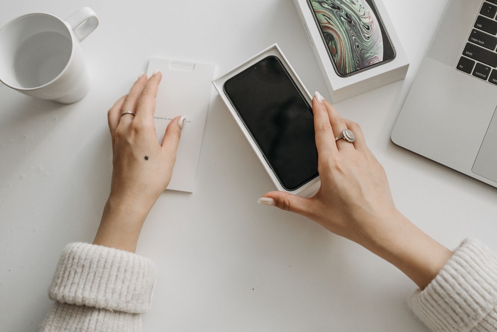 Hands unboxing a new iPhone on a desk beside a laptop and a coffee mug