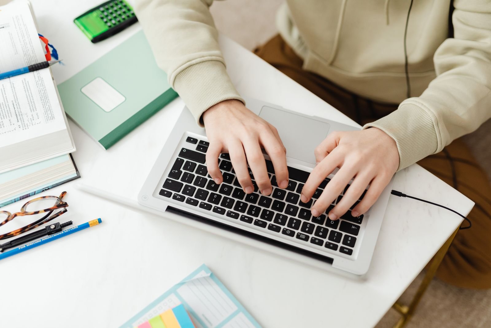 Overhead view of hands typing on a white laptop surrounded by study books and stationery