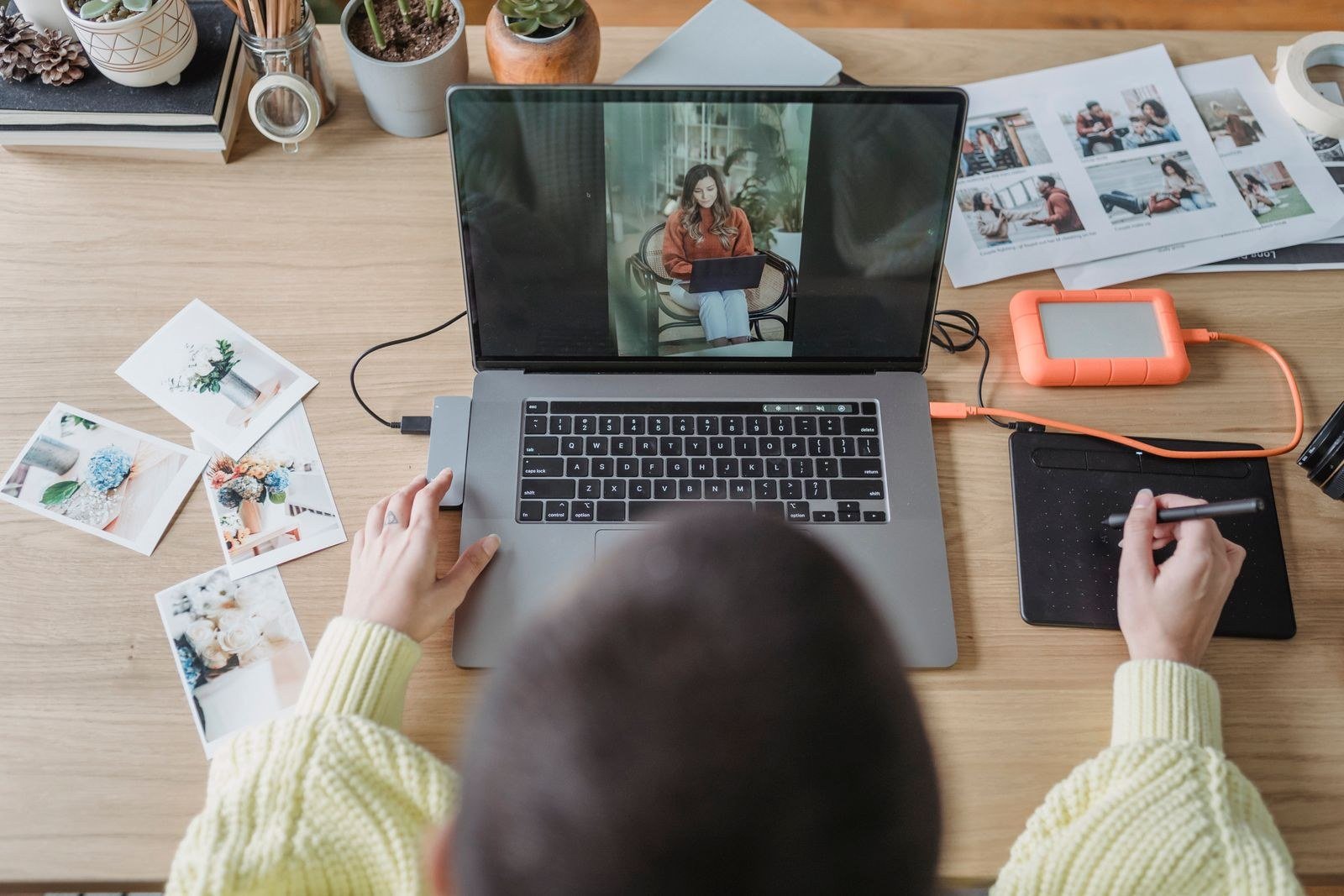Overhead view of a photographer drawing on a graphics tablet while editing photos on a laptop, with printed photos on the table