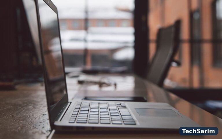 A laptop in profile on a long wooden desk in front of large office windows