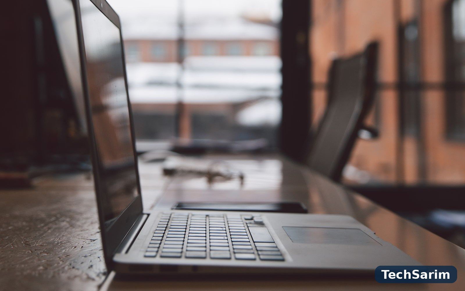 A laptop in profile on a long wooden desk in front of large office windows