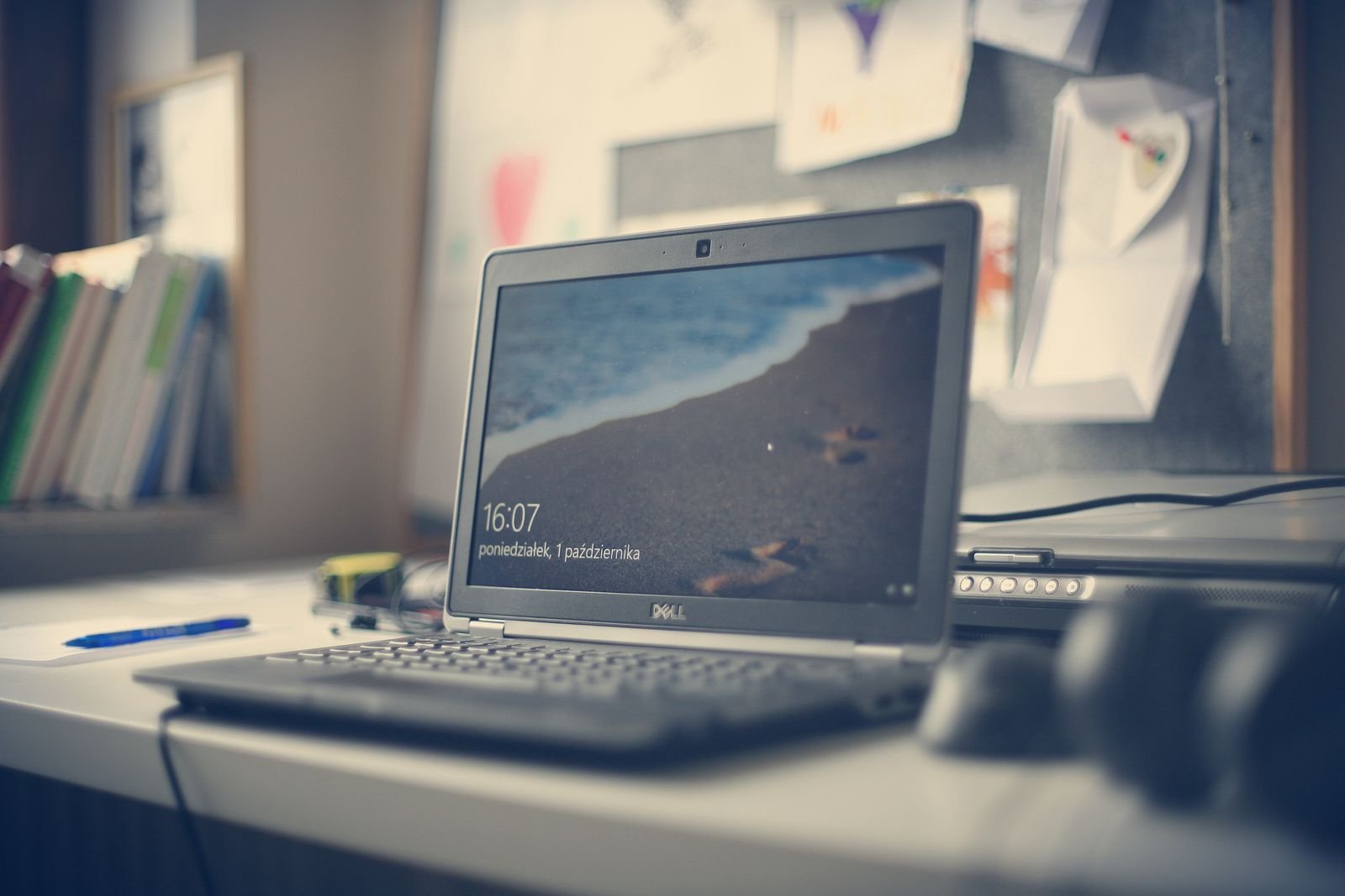 A laptop on a desk showing its lock screen with a beach wallpaper, with books and a pinboard behind