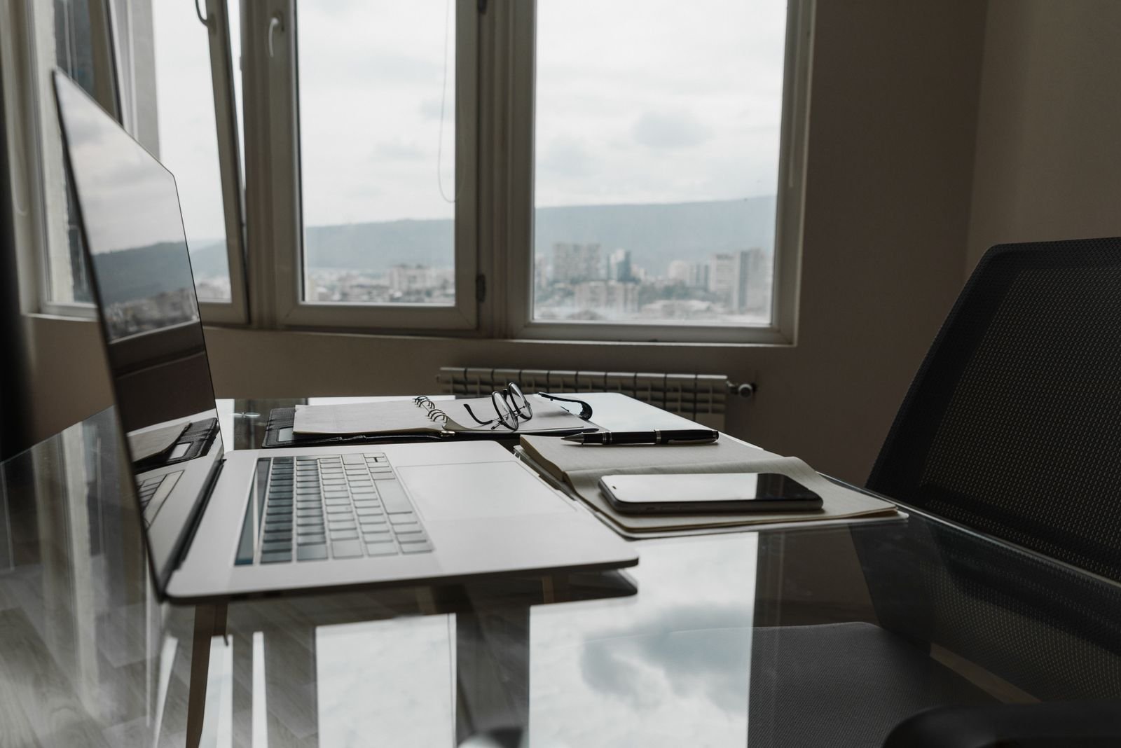 A laptop on a glass desk beside a notebook, glasses and smartphone, with a city view through the window behind