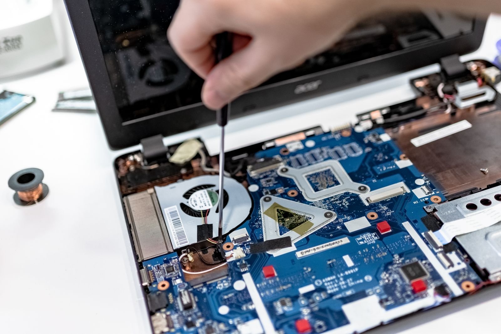 A hand driving a screw inside an opened laptop, beside the cooling fan and motherboard