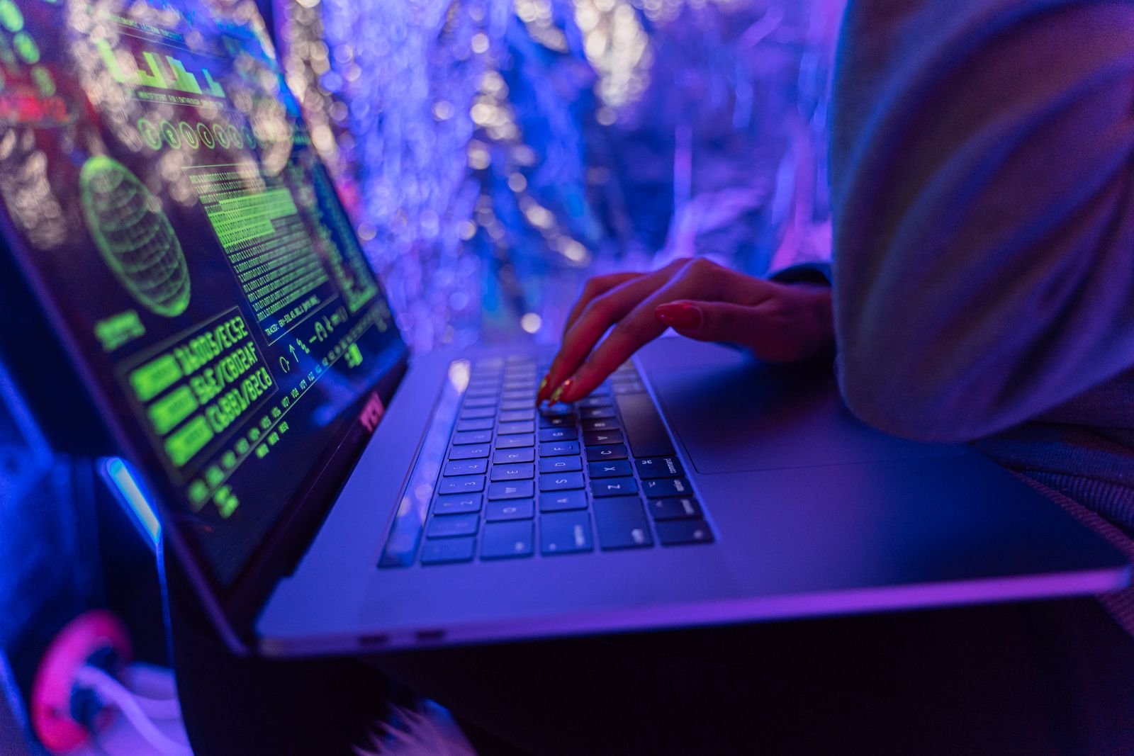 Close-up of a woman's hand with red nail polish typing on a laptop showing green security data graphics, lit by purple light