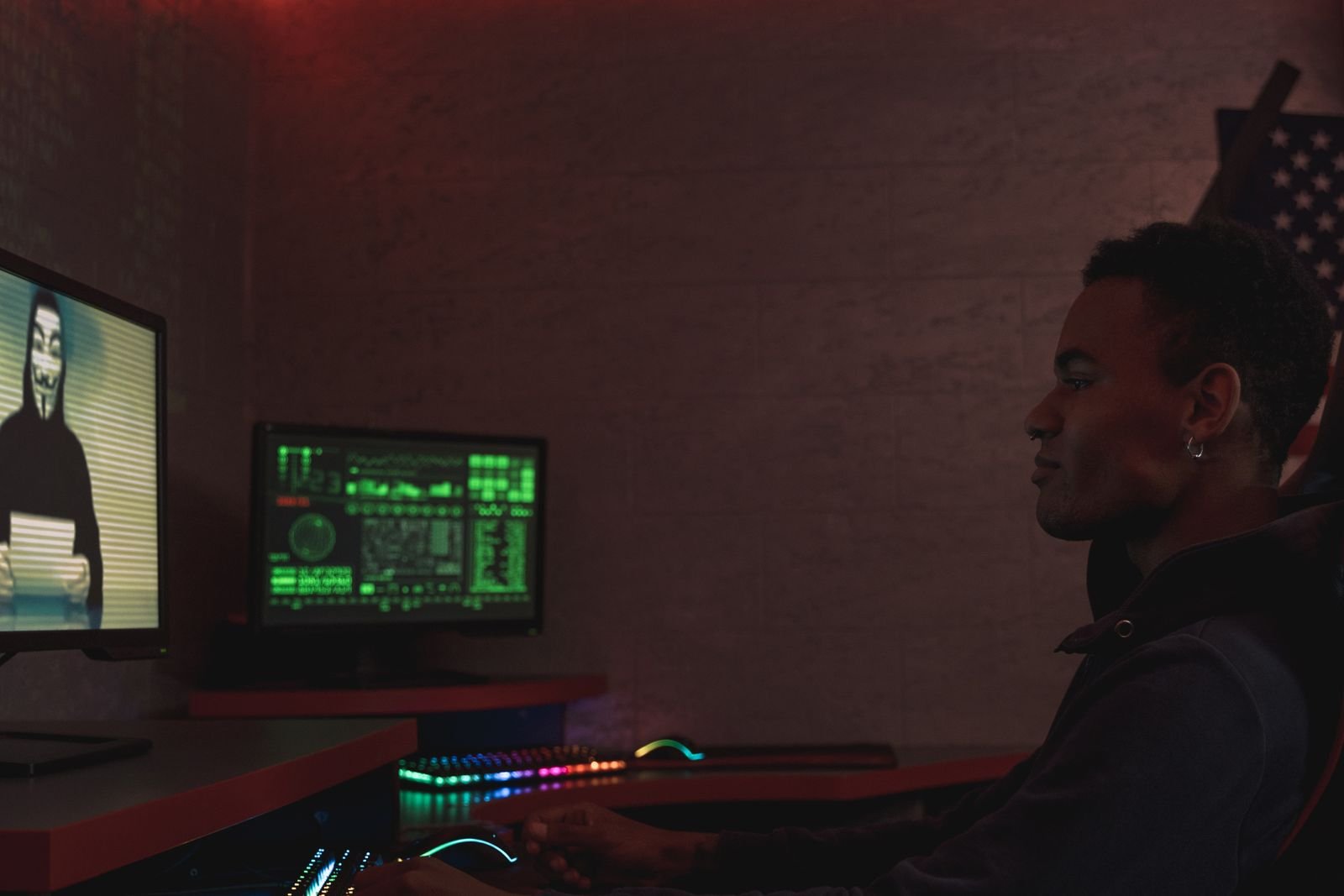 A man in a dark room sitting at a desk with two monitors, one showing green data dashboards and the other a masked figure on screen