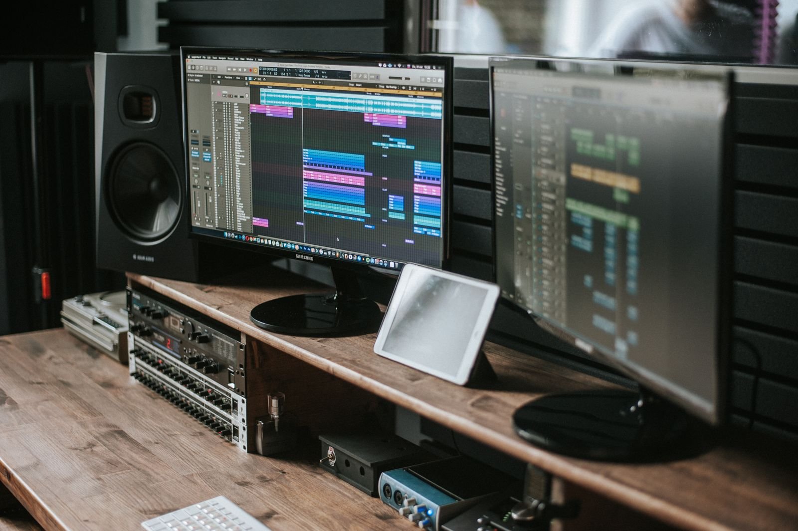 A studio desk with two monitors showing editing timeline software, a studio monitor speaker and audio mixing gear on a wooden desk