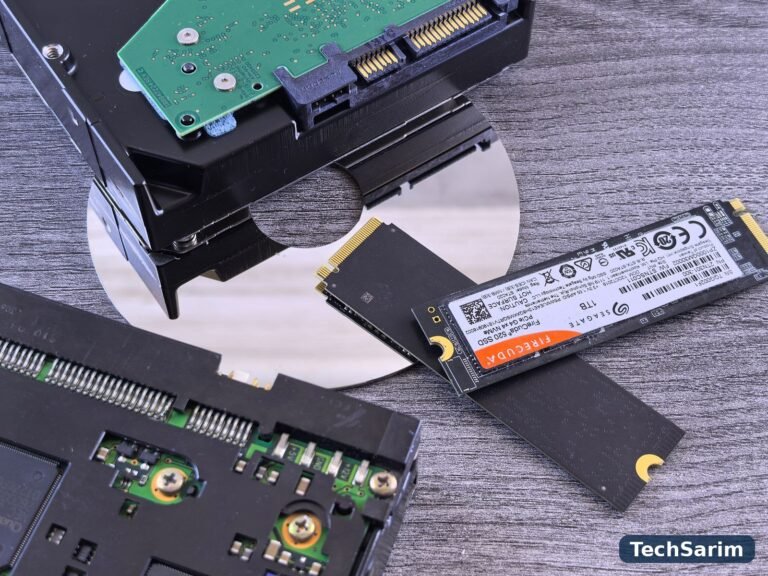 A hard disk drive with its platter exposed beside two M.2 NVMe SSD sticks on a grey wooden surface