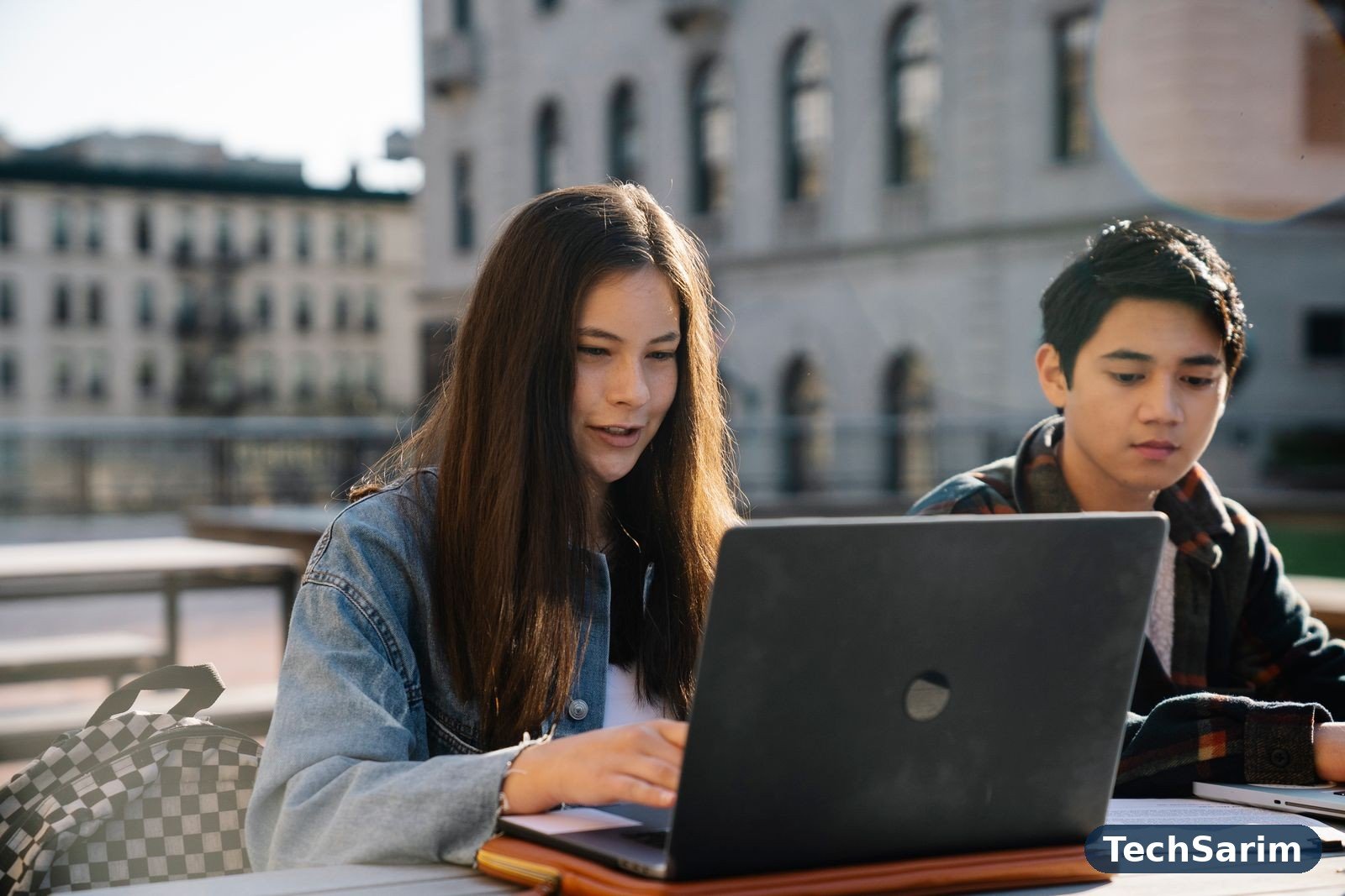 Two students working together on a laptop at an outdoor table on campus, with a historic university building behind them