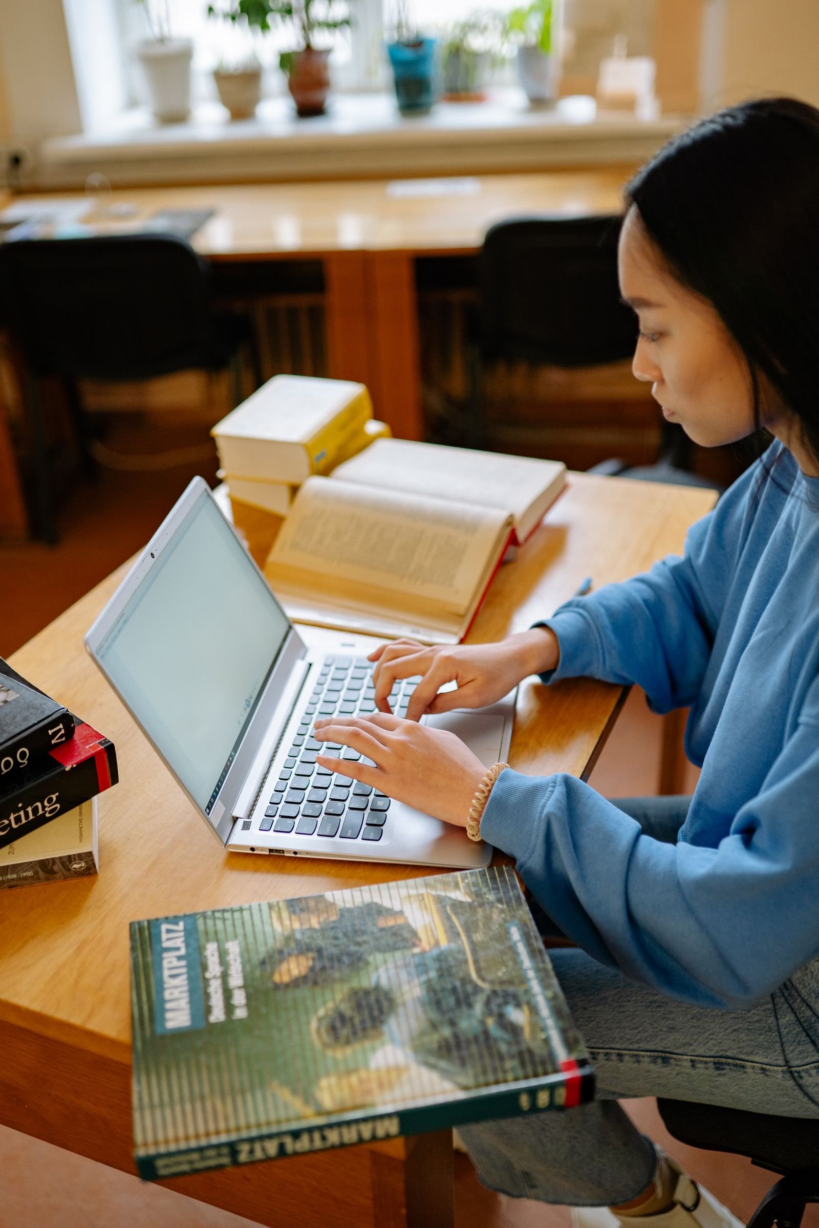 A student typing on a slim laptop at a wooden library desk, surrounded by open textbooks