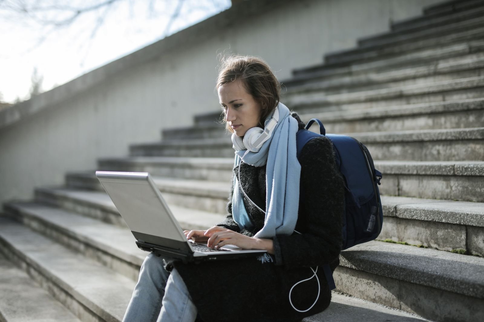 A student with headphones around her neck and a backpack beside her, working on a laptop while sitting on outdoor campus steps