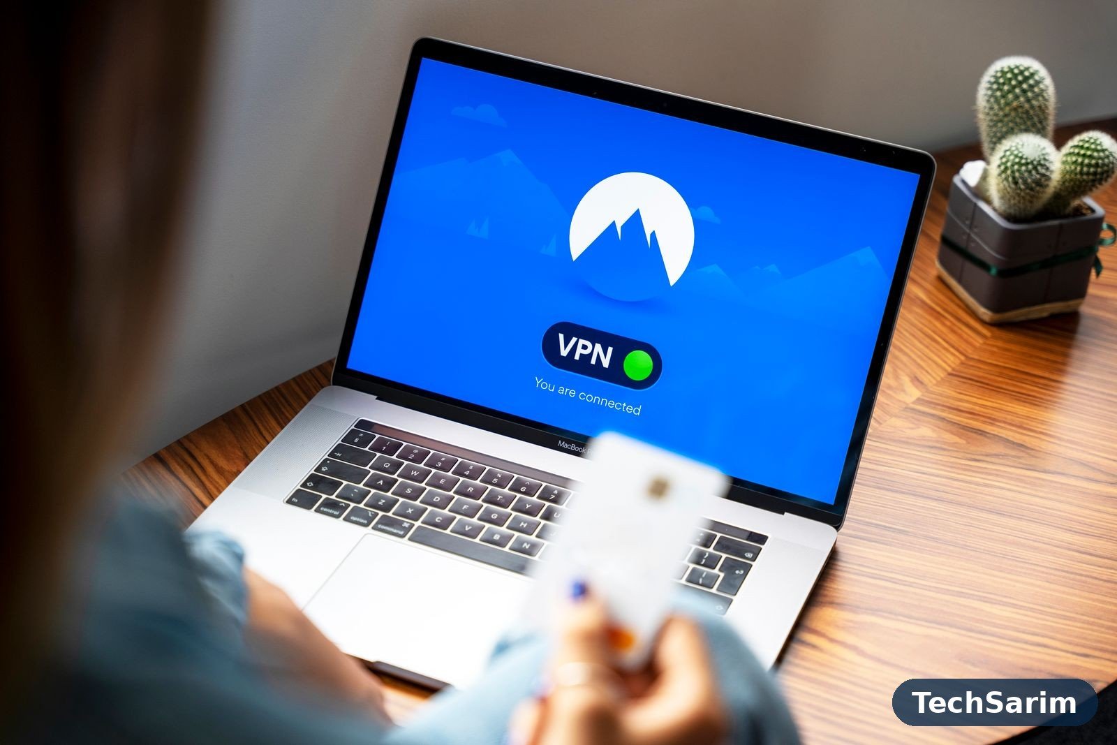 A laptop on a wooden desk displaying a VPN app's blue connected screen, with the user holding a payment card in the foreground and a small potted cactus beside the laptop