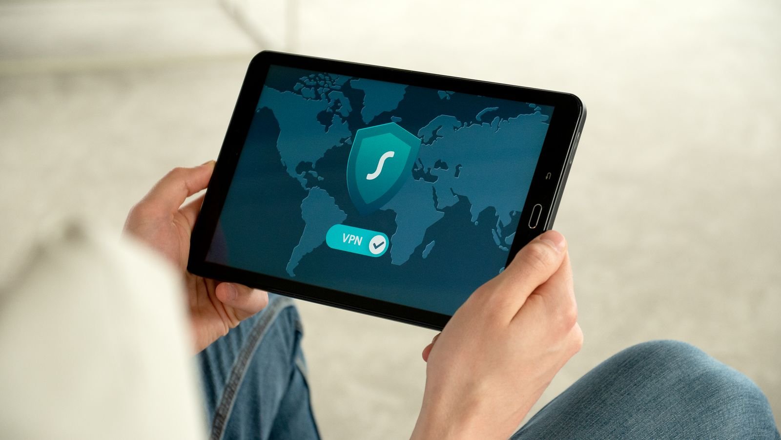 Hands holding a tablet whose screen shows a VPN app marked connected, with a shield icon over a world map