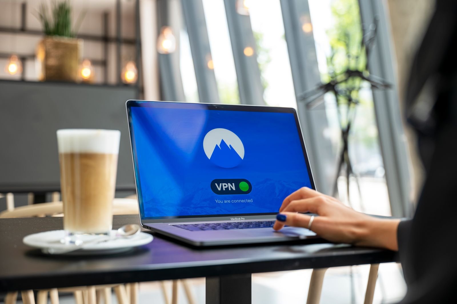 A laptop on a café table showing a VPN app's connected screen, with a latte on a saucer beside it