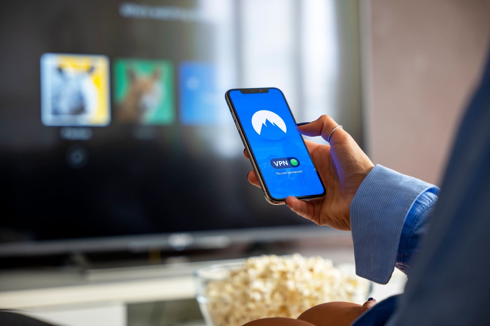 A hand holding a smartphone showing a VPN app connected, with a television's streaming menu blurred behind it and a bowl of popcorn below