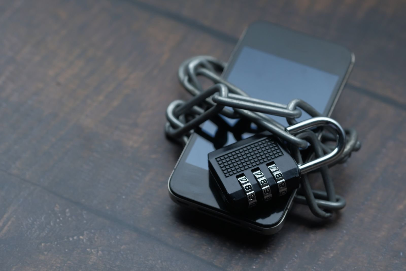 A smartphone wrapped in a heavy metal chain and secured with a black combination padlock on a dark wooden surface