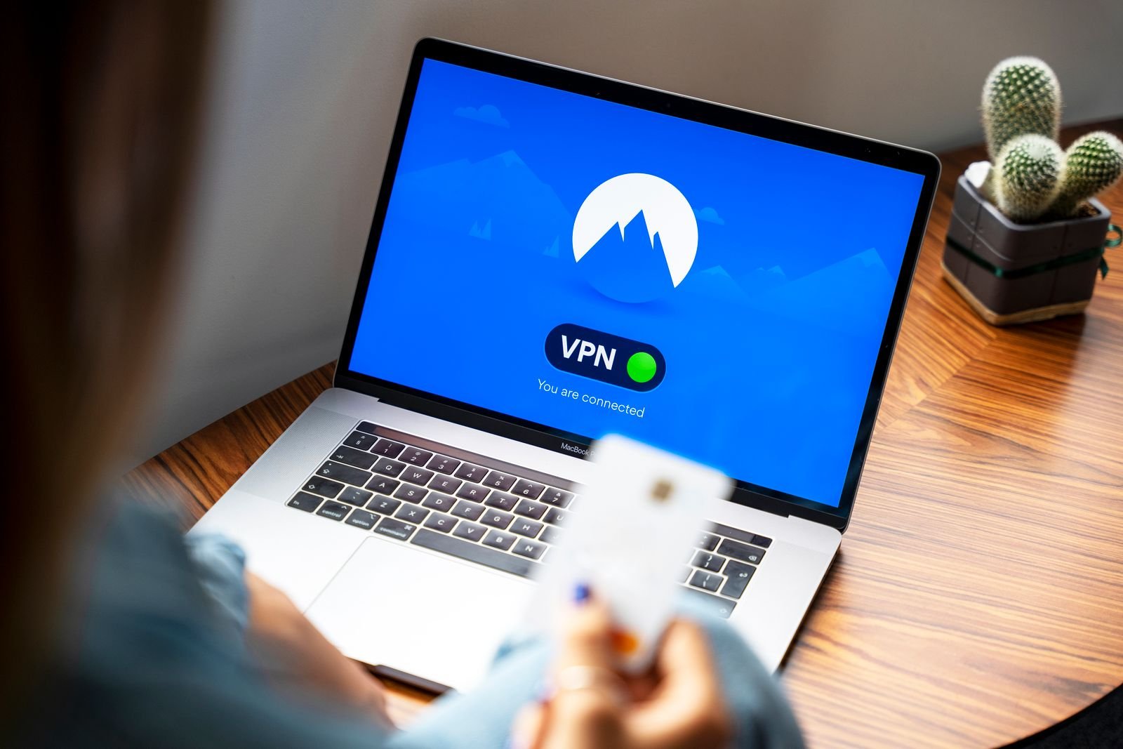 A laptop on a wooden desk showing a VPN app with a green connected indicator, a person's hands holding a payment card blurred in the foreground and a small potted cactus beside the laptop