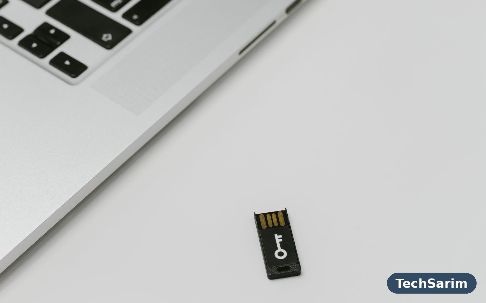 A silver laptop on a white desk with a small black USB security key bearing a white key symbol lying beside it