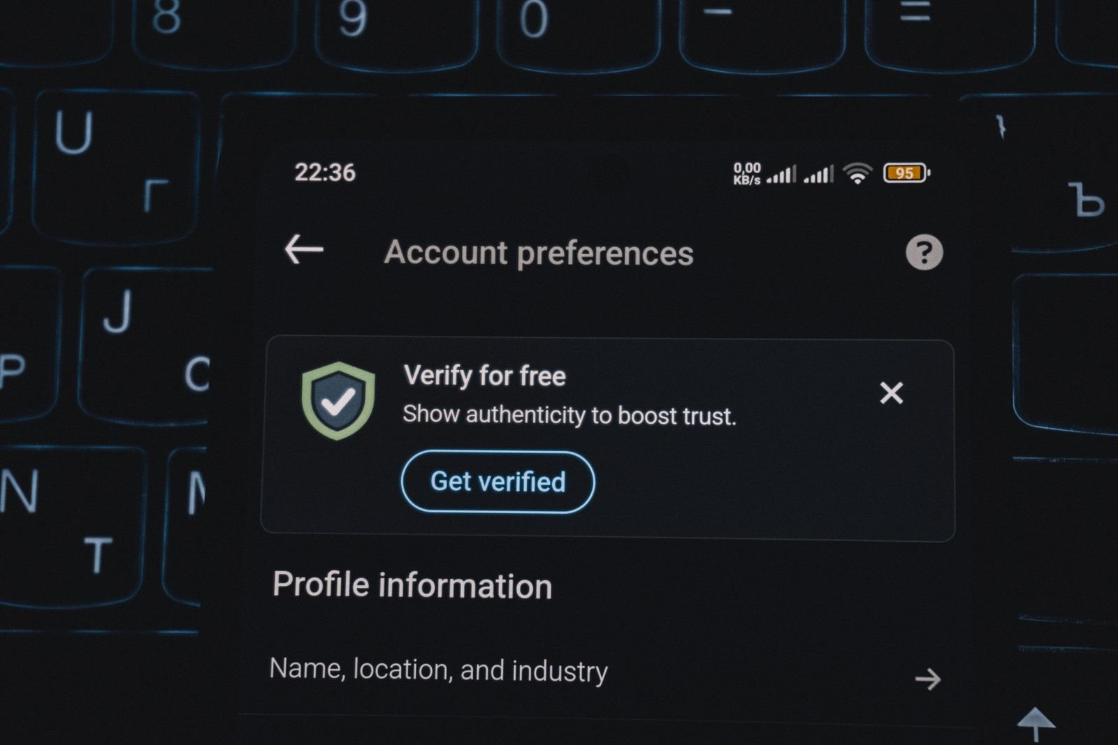 Close-up of a smartphone screen showing an account preferences page with a verification prompt and shield icon, resting on a backlit laptop keyboard