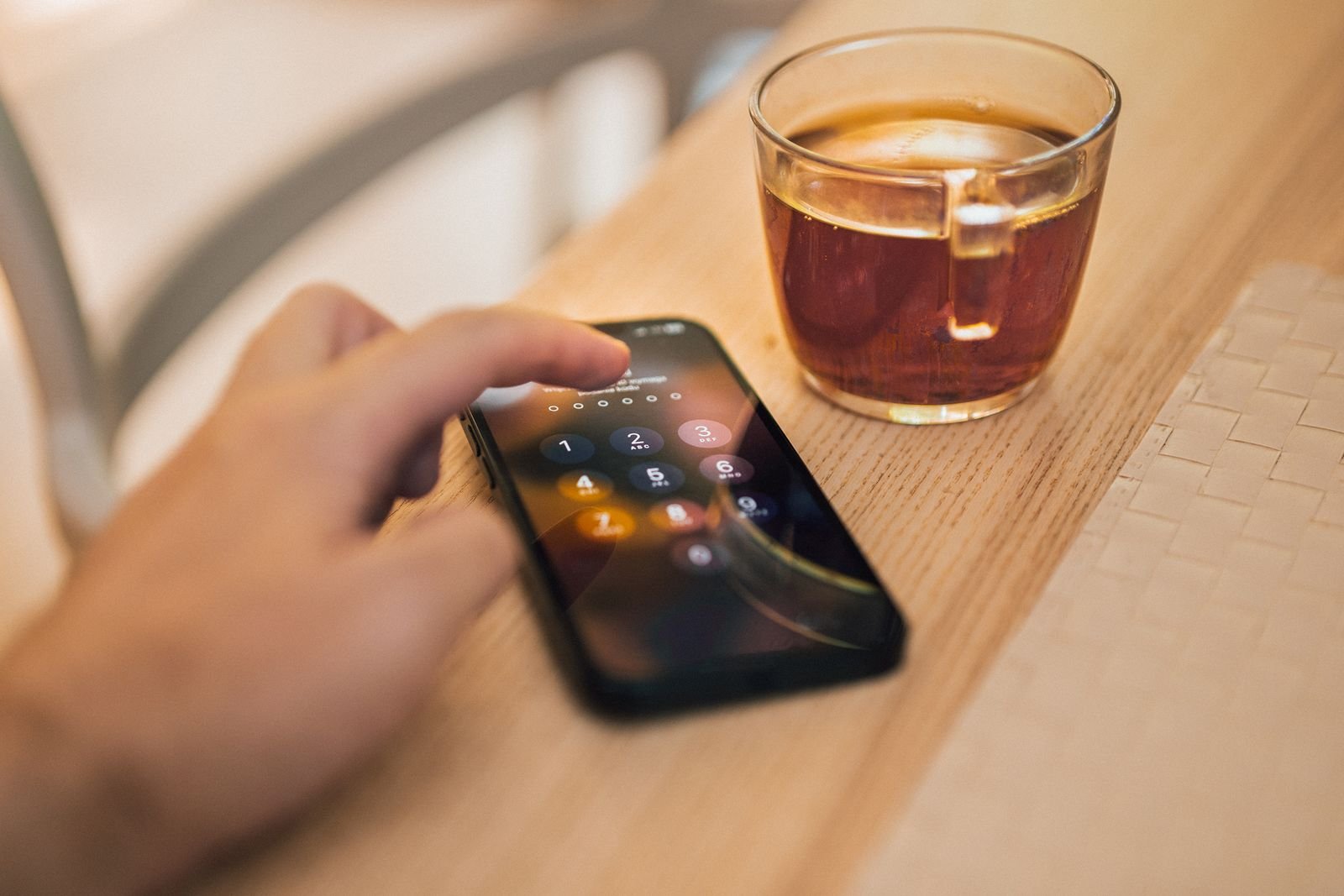 A hand unlocking a smartphone with a passcode on a wooden table beside a glass cup of tea