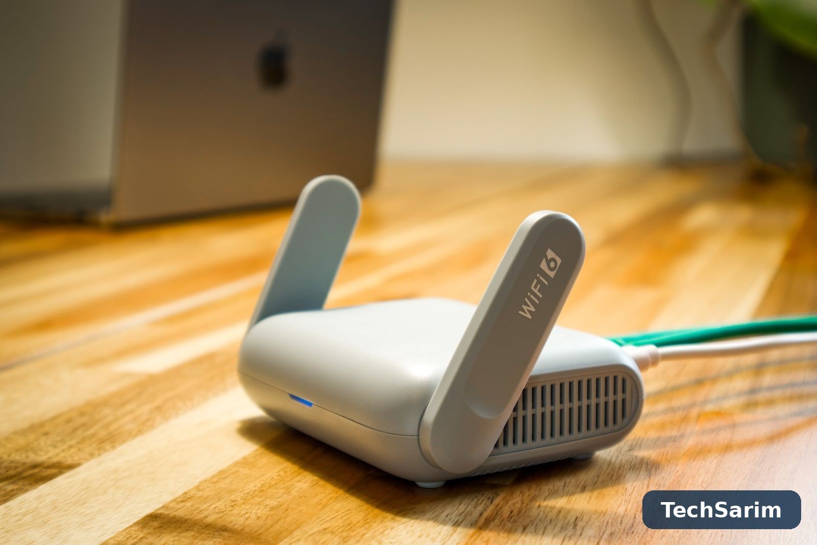 A compact grey WiFi 6 router with two paddle antennas and a glowing blue status light on a wooden desk, with a laptop blurred behind it