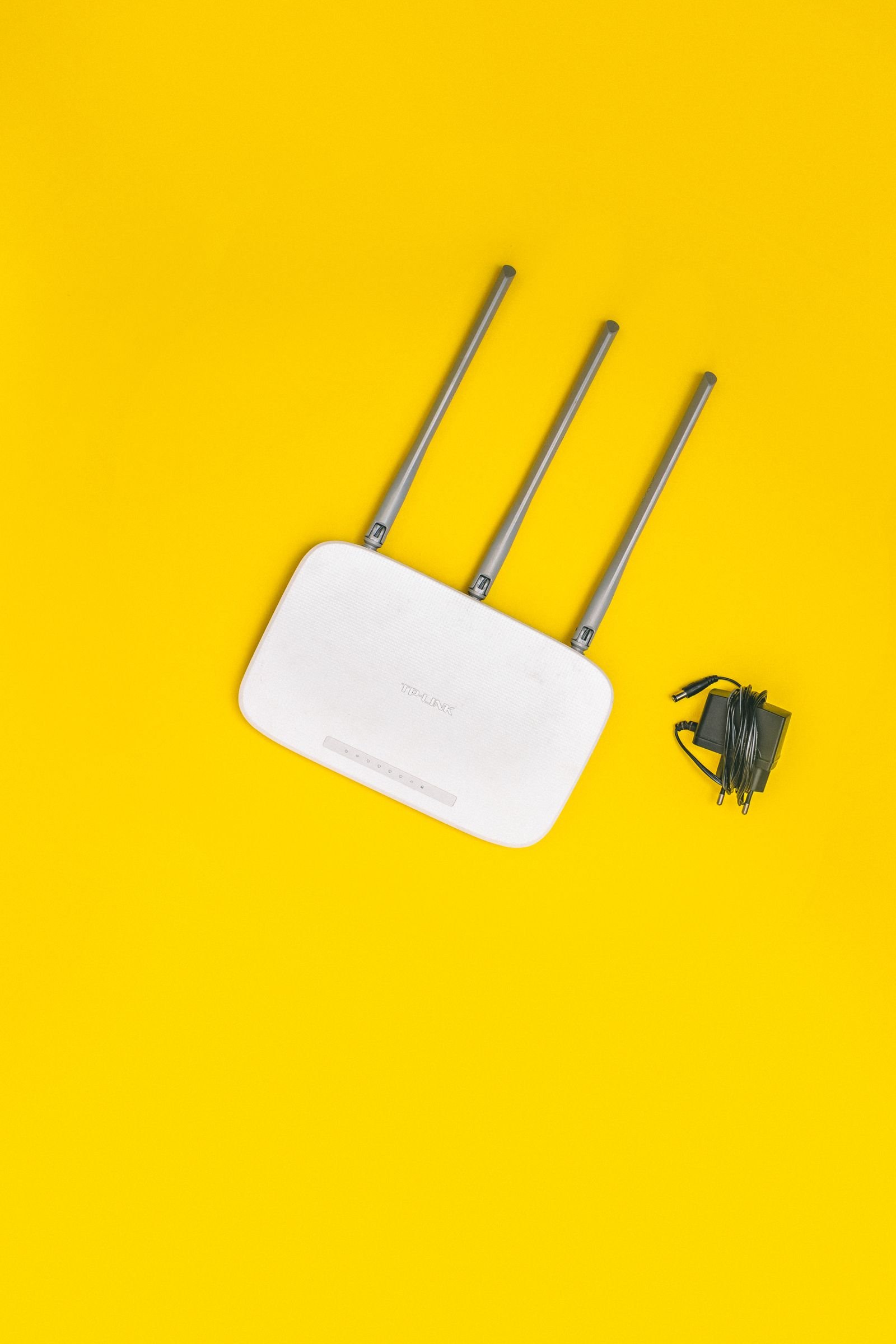 A white router with three tall antennas and its power adapter on a bright yellow background, photographed from above
