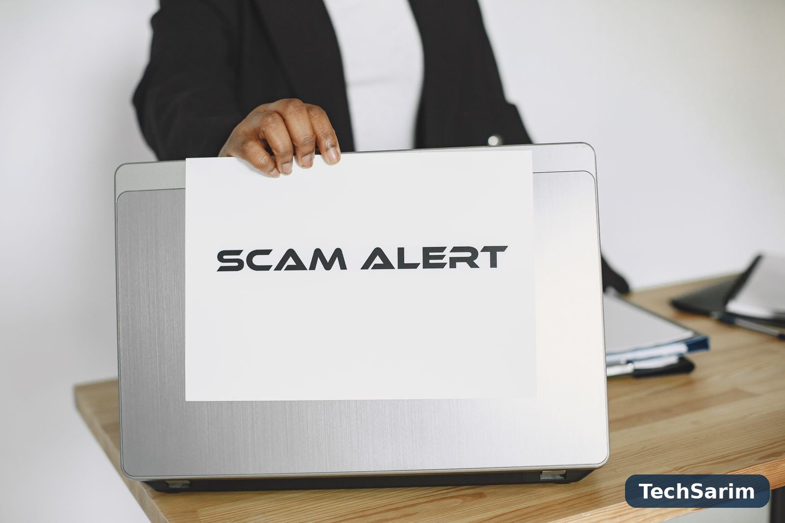 A person in a suit jacket holding a white sign reading SCAM ALERT in front of a silver laptop on a wooden desk
