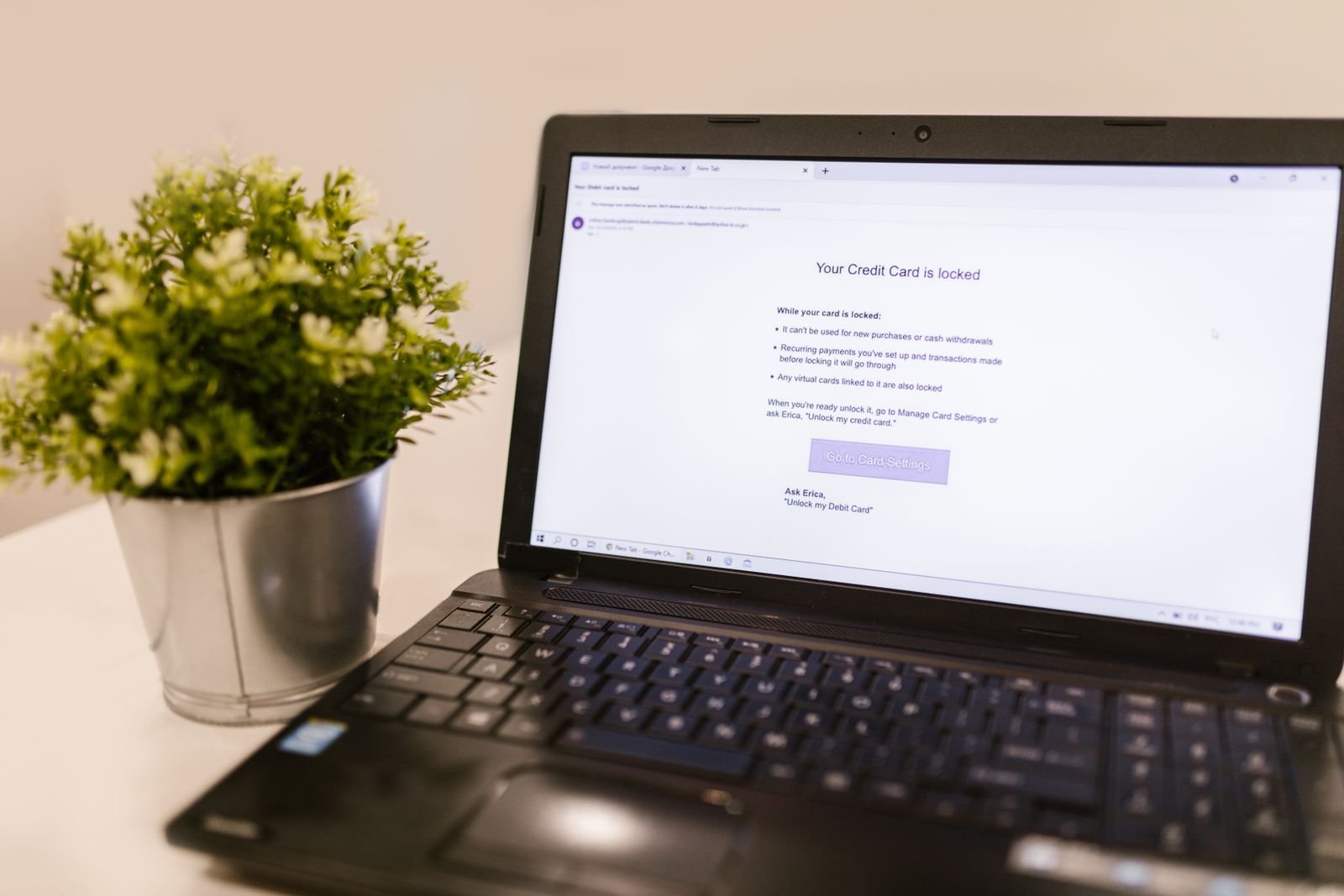 A laptop on a white desk beside a potted plant, its screen showing a bank notification page saying a credit card is locked