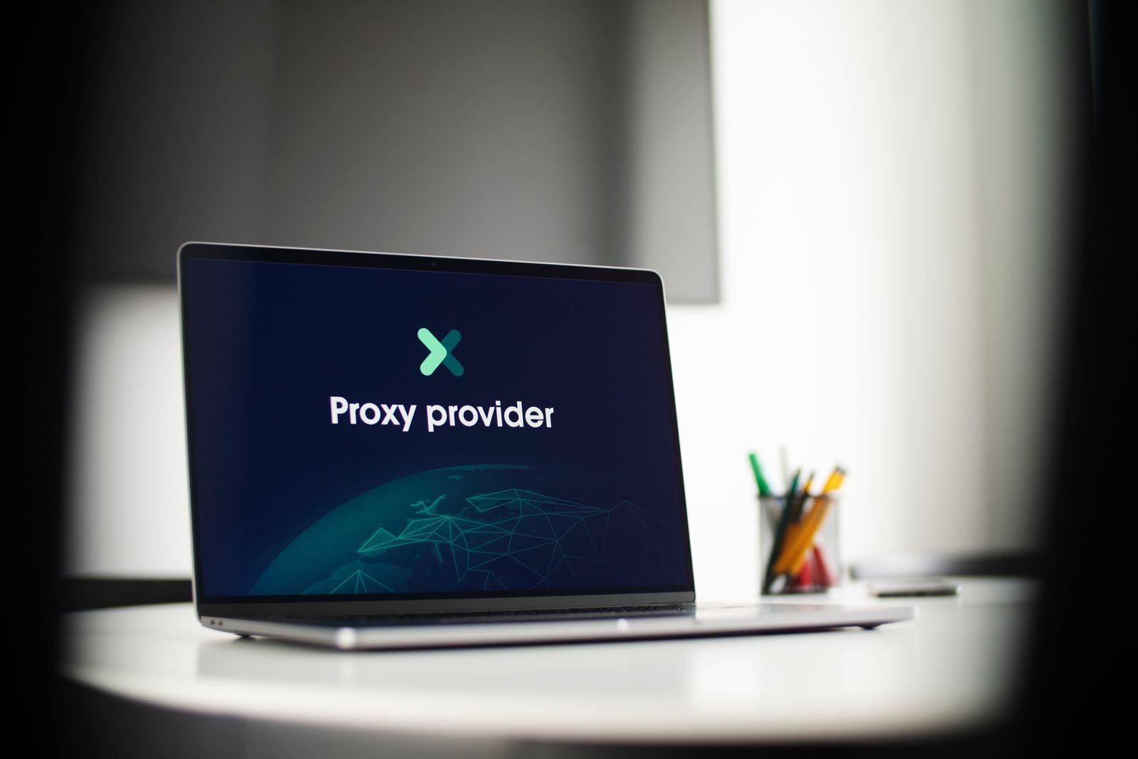 A laptop on a desk in a dim office displaying the words 'Proxy provider' above a wireframe globe graphic, with a pot of pencils behind it