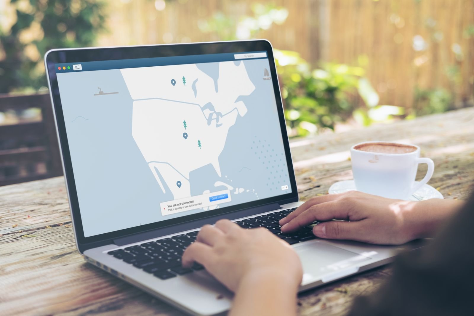 Hands typing on a laptop at an outdoor wooden table, the VPN app's map on screen showing 'You are not connected', a cup of coffee on a saucer beside it