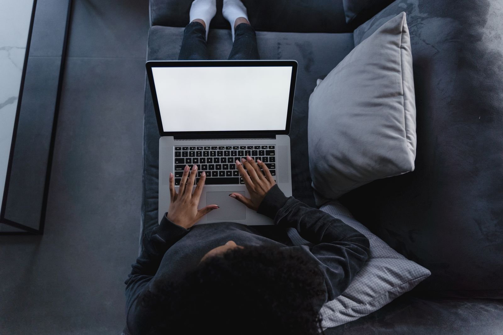 Overhead view of a person lying on a dark grey sofa at home, typing on a laptop balanced on their lap