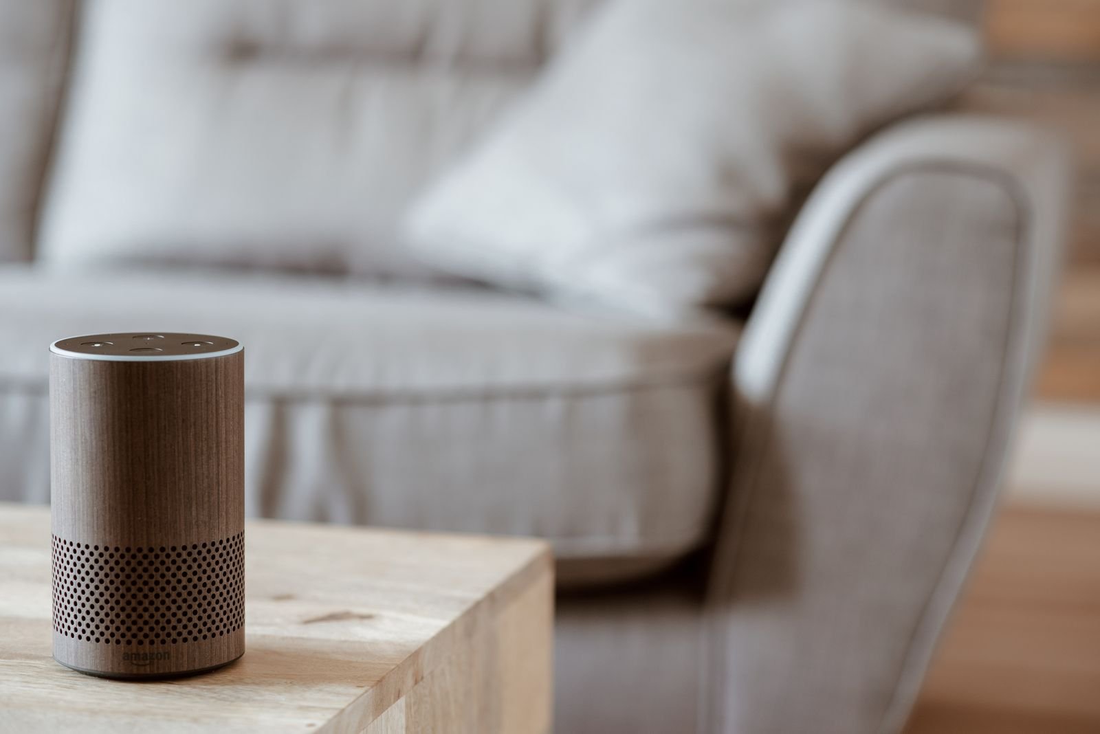 A wood-finish Amazon Echo smart speaker on a wooden side table in front of a grey sofa in a living room