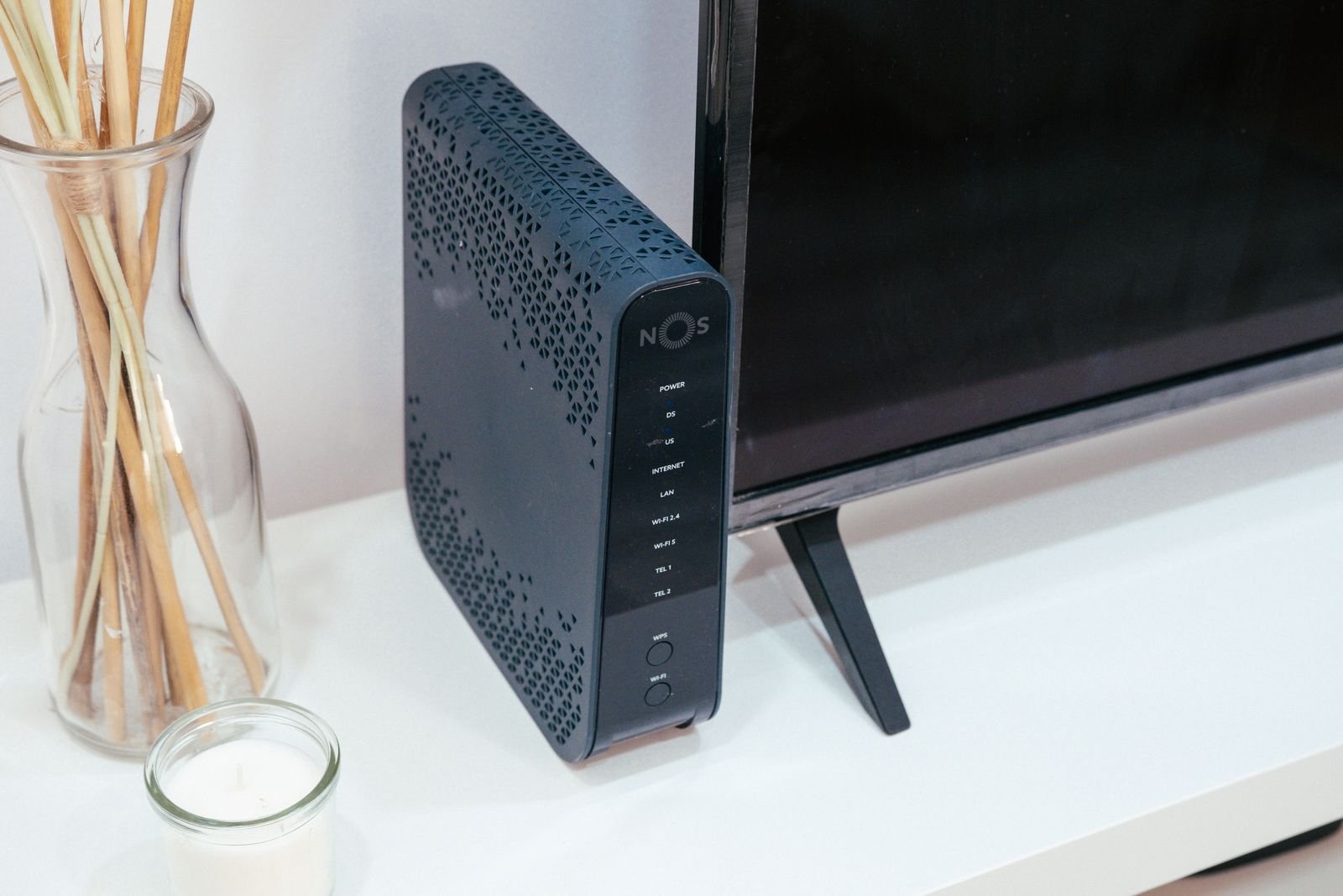 A black upright ISP router standing on a white TV stand beside a television, its status labels and WPS button visible on the front edge
