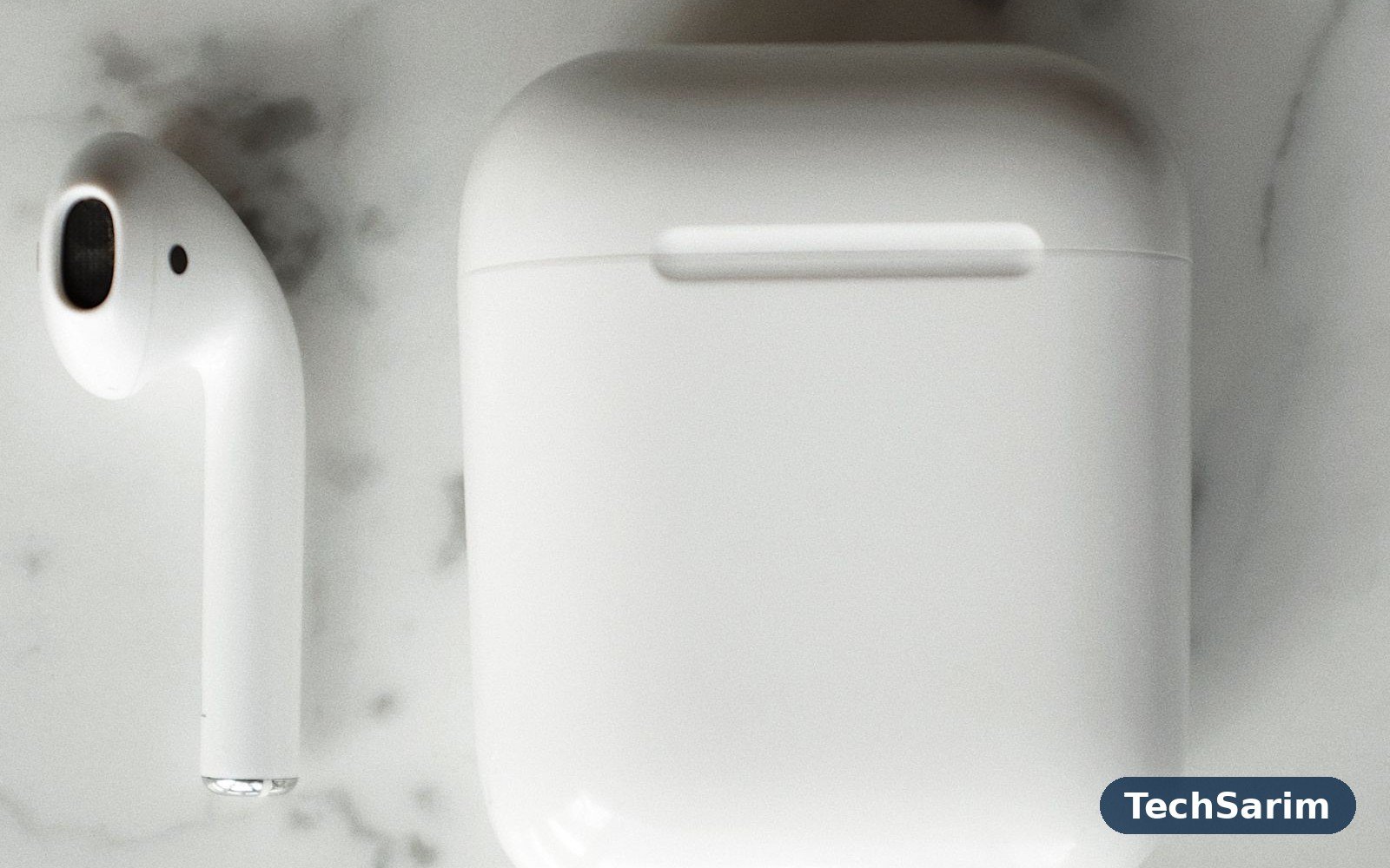 Top-down view of a white wireless earbud beside its closed white charging case on a grey marble surface