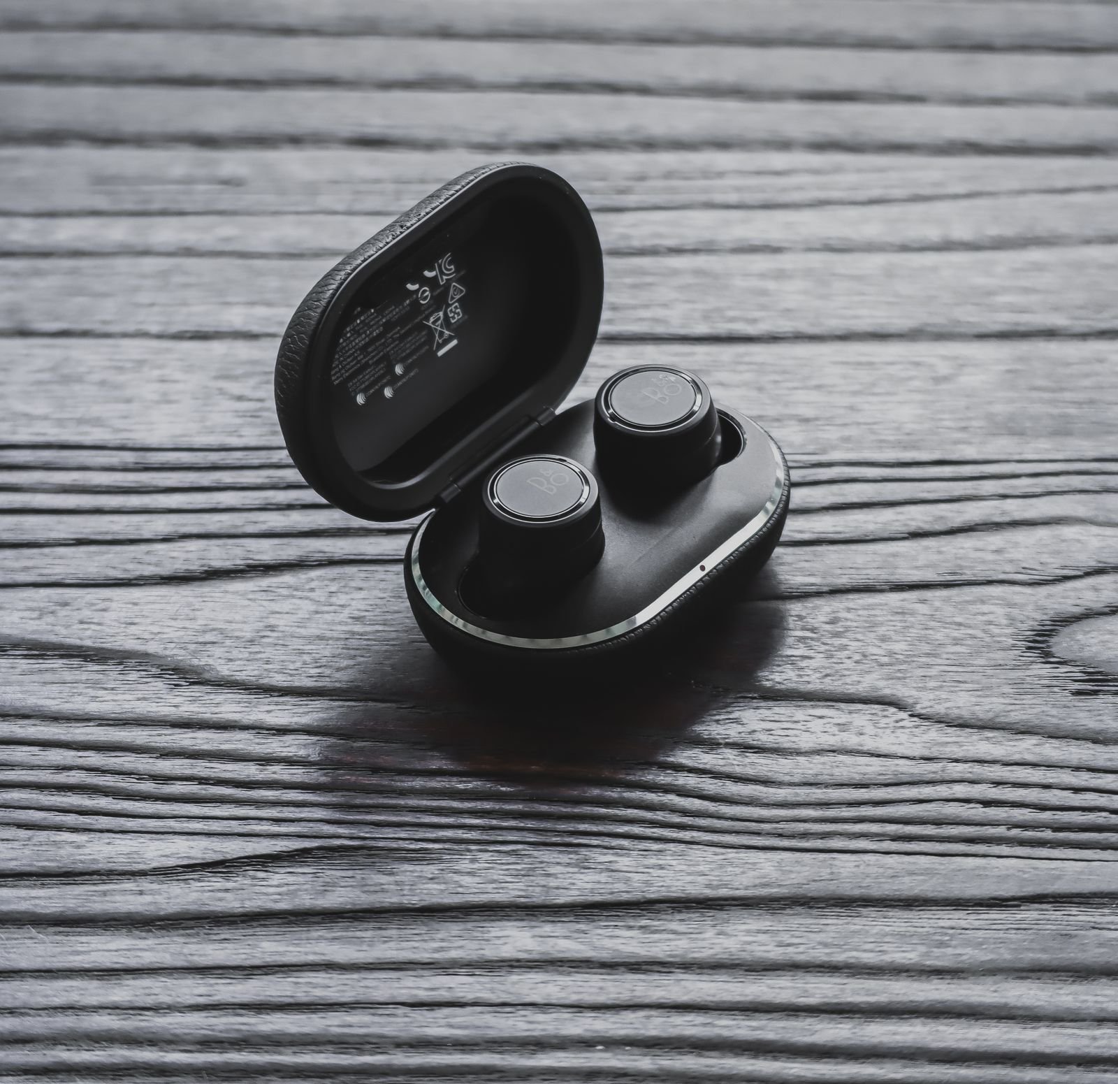 Two round black wireless earbuds sitting in an open black leather charging case on a dark wooden table