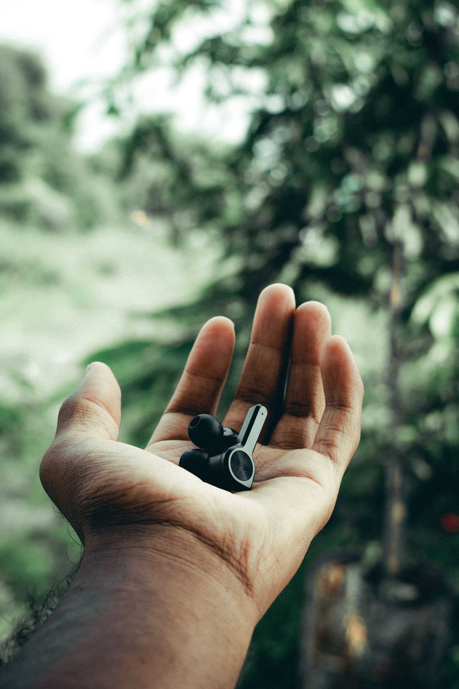 An open hand holding a pair of black stem-style wireless earbuds against blurred green foliage outdoors