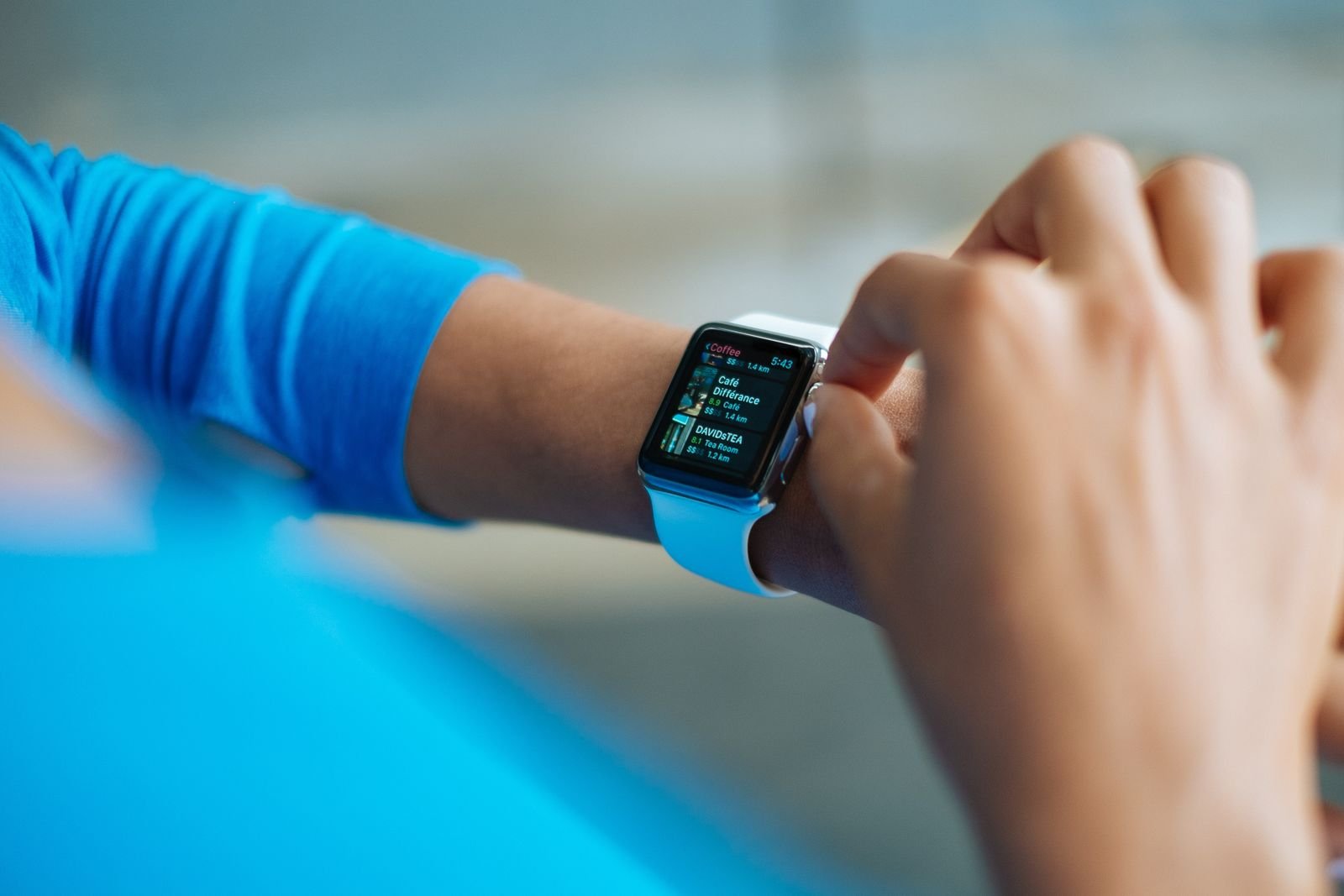 A person in a blue top scrolling through a list of nearby coffee shops on a rectangular smartwatch with a white strap