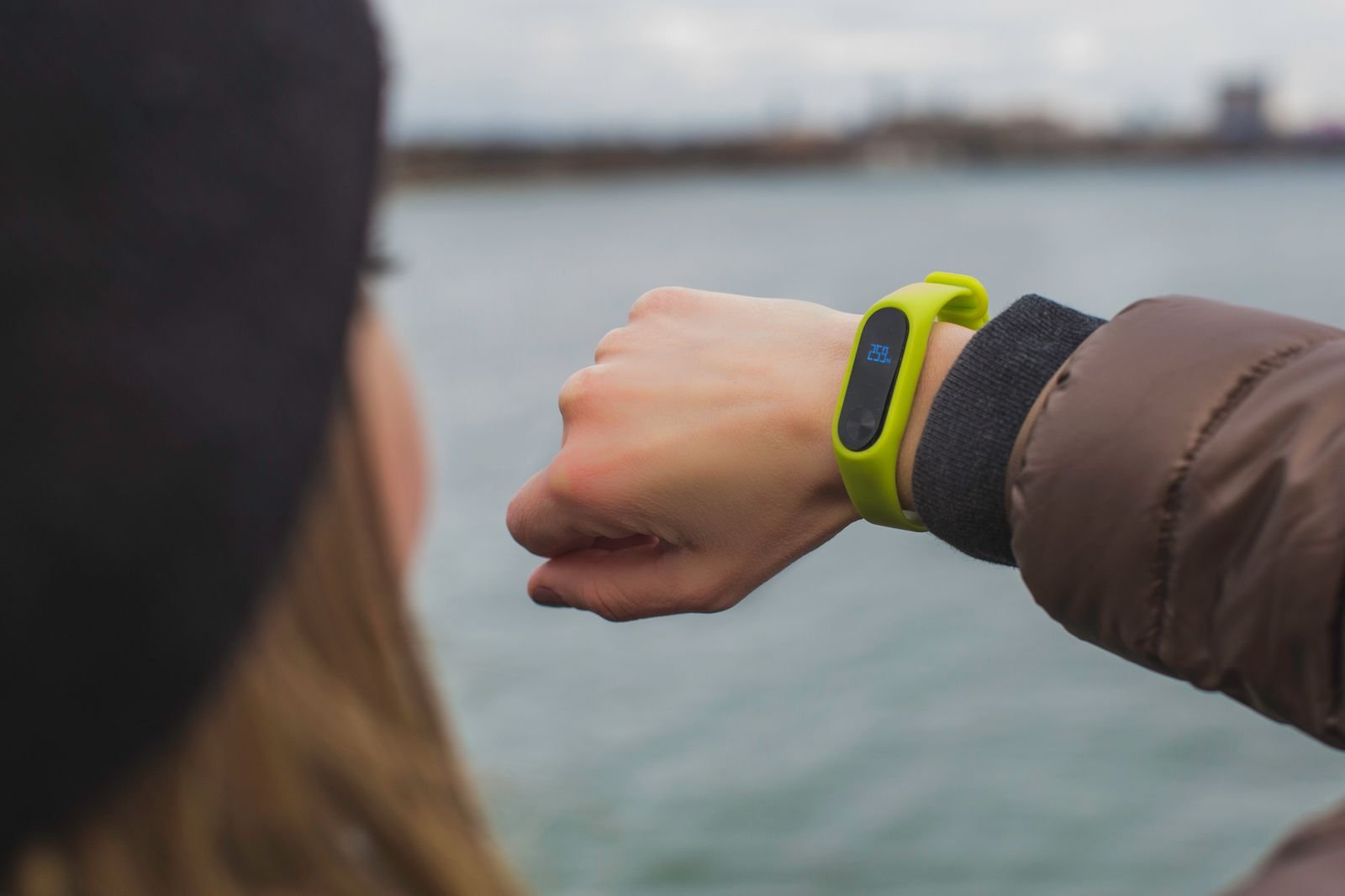 A woman outdoors by a lake checking a slim lime-green fitness band on her wrist, its small screen showing a step count
