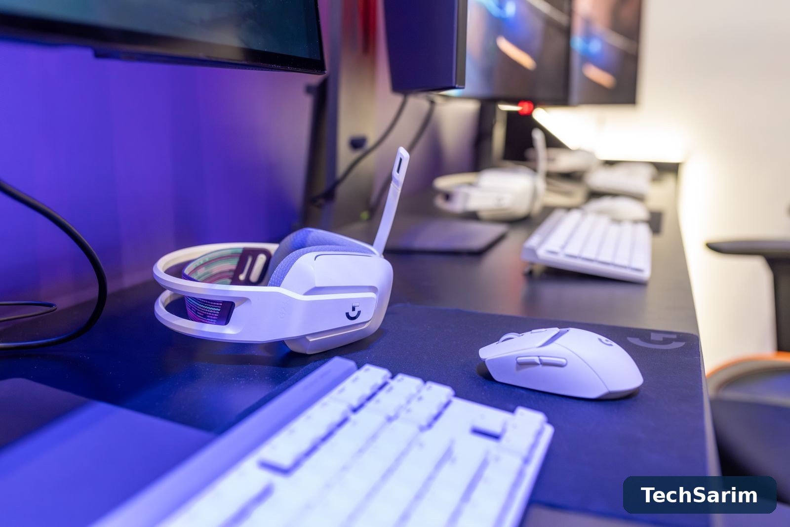 A white gaming headset and white wireless mouse resting on a large dark desk mat, with a blurred white keyboard in the foreground and monitors behind, lit in purple and blue