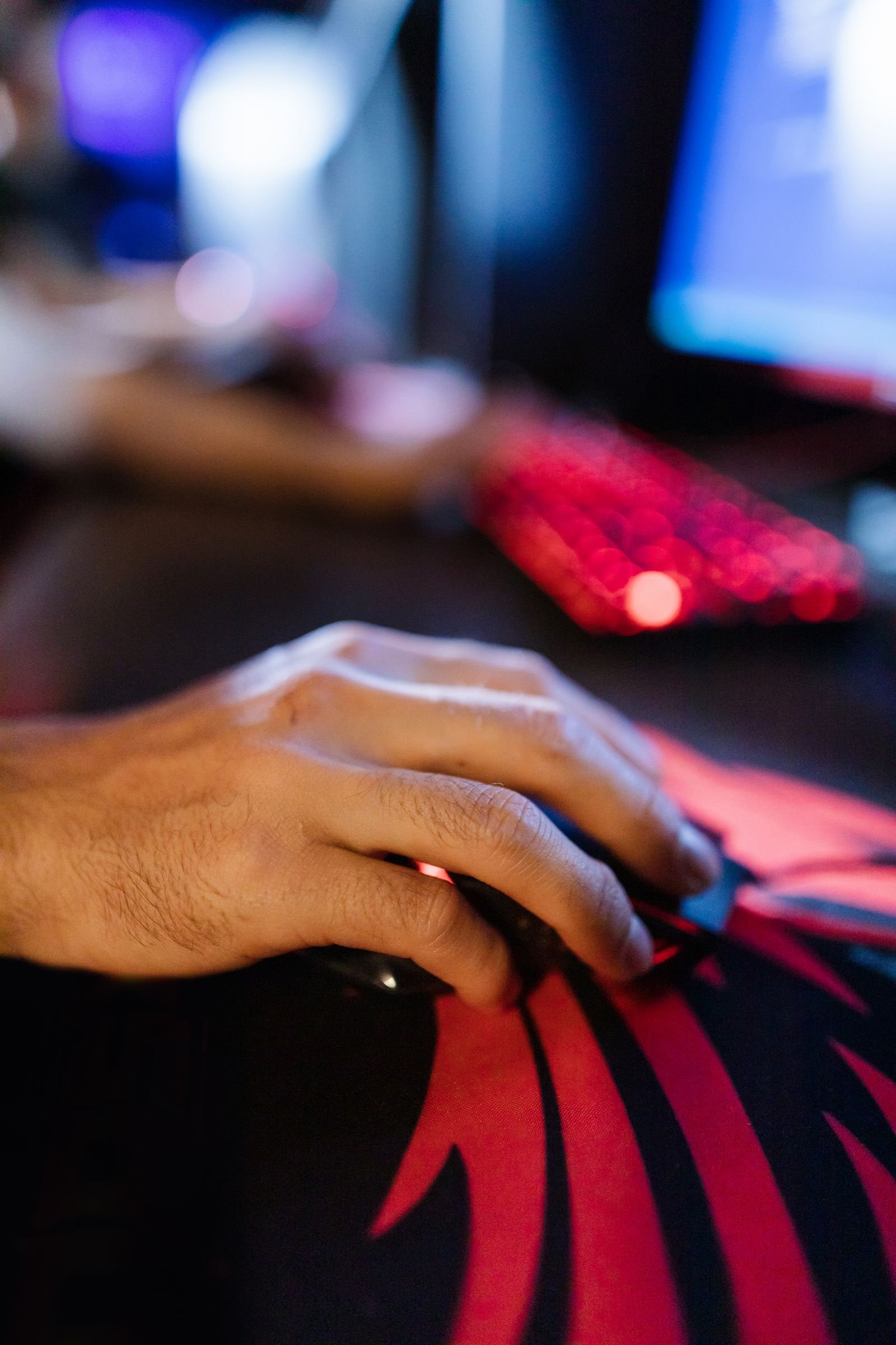 Close-up of a hand gripping a gaming mouse on a red and black patterned mouse pad, with a red-backlit keyboard blurred in the background