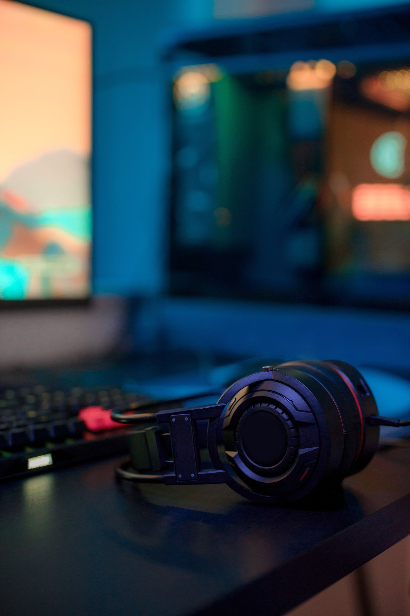 Close-up of a black gaming headset lying on a desk in front of a blurred monitor and a glowing PC case, with part of a keyboard visible beside it