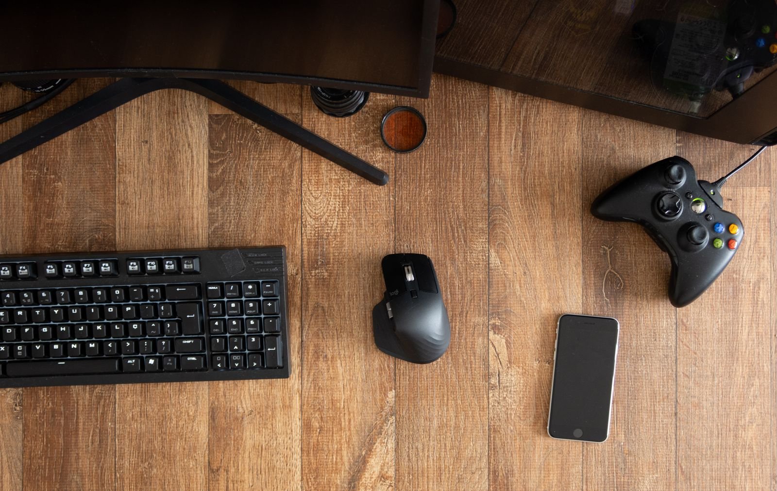 Top-down view of a black full-size keyboard, a black wireless mouse, a game controller and a smartphone arranged on a wooden desk
