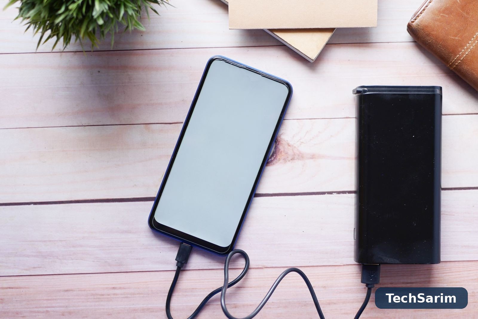 Top-down view of a smartphone charging by cable from a black power bank on a light wooden desk, with a notebook and wallet nearby