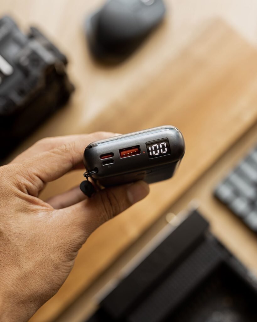 Close-up of a hand holding a compact power bank showing its USB-C and USB-A ports and an LED battery display reading 100 percent
