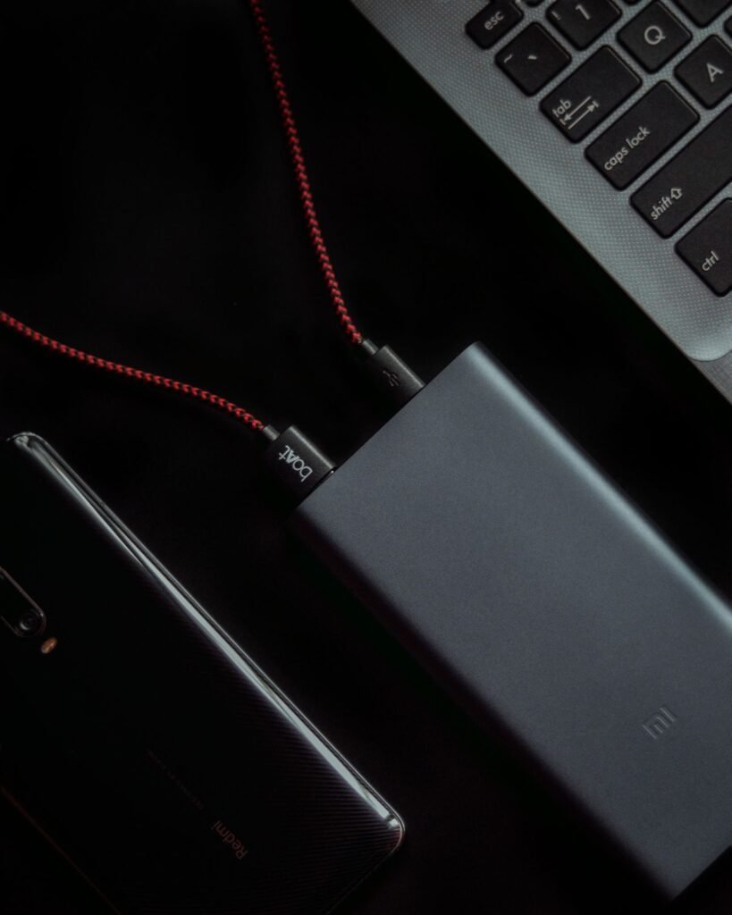 A grey metal power bank connected with red braided cables beside a smartphone and the corner of a laptop keyboard on a dark desk