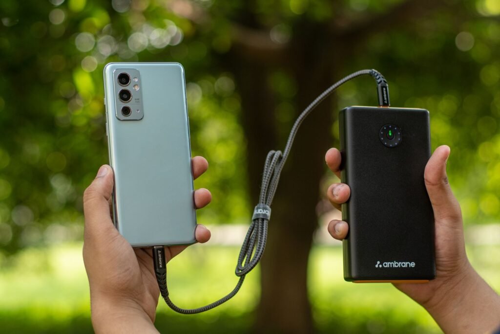 Two hands holding a smartphone and a large black power bank connected by a braided cable outdoors, with green trees behind